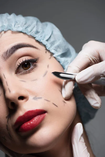 Close up of plastic surgeon in latex gloves holding scalpel near woman with and marked lines on face isolated on grey — Stock Photo