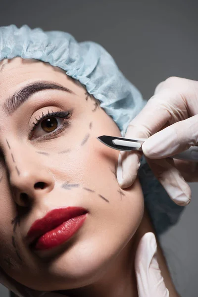 Primer plano del cirujano plástico en guantes de látex que sostienen el bisturí cerca de la mujer con y líneas marcadas en la cara aislada en gris - foto de stock