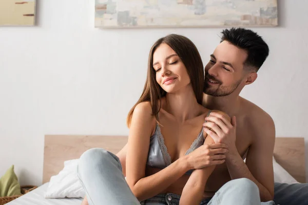 Hombre feliz abrazando a mujer seductora sonriendo con los ojos cerrados en la cama - foto de stock