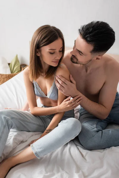 Sexy young man hugging pretty woman sitting on bed in jeans and bra — Stock Photo