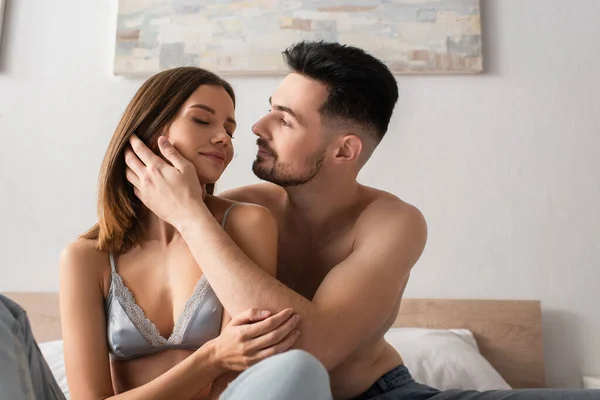 Без сорочки чоловік торкається обличчя сексуальної дівчини, сидячи на ліжку з закритими очима — стокове фото