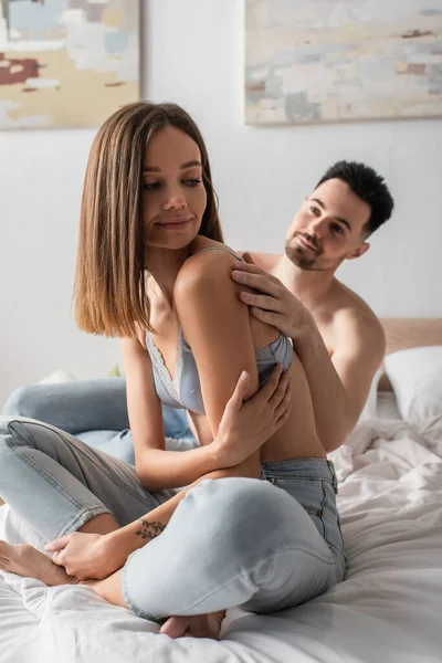 Сексуальний чоловік спокушає красиву молоду жінку, сидячи на ліжку в джинсах і бюстгальтері — стокове фото