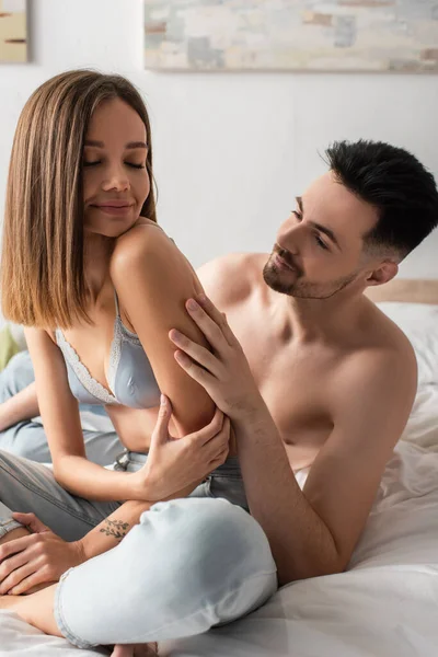 Hombre sin camisa abrazando a mujer sonriente en sujetador y jeans sentados en la cama con las piernas cruzadas - foto de stock