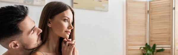 Young man near sensual dreamy woman looking away in bedroom, banner — стоковое фото