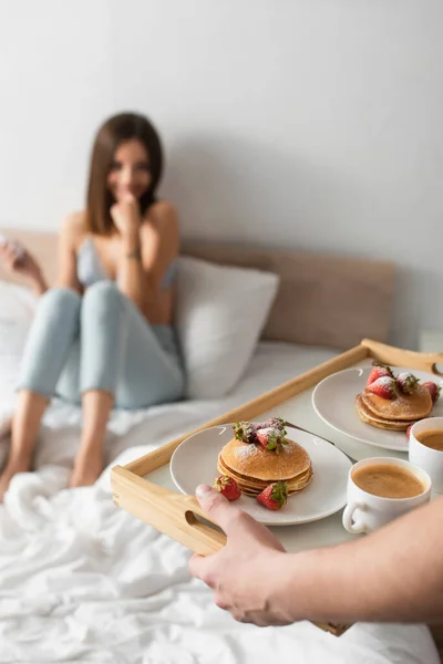 Selective focus of tasty pancakes with strawberries on tray in hands of man near blurred woman in bedroom — Stock Photo