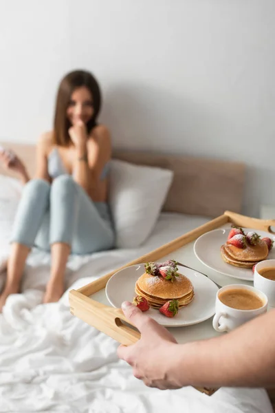 El foco selectivo de los crepes sabrosos con las fresas en la bandeja en las manos del hombre cerca de la mujer borrosa en el dormitorio - foto de stock