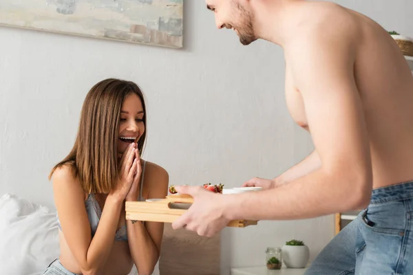 Shirtless man holding tray with delicious breakfast near amazed girlfriend in bedroom — Stock Photo