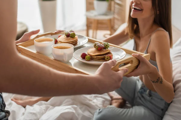 Vista recortada de hombre sosteniendo bandeja con panqueques y café cerca de la mujer sexy y feliz en el dormitorio - foto de stock