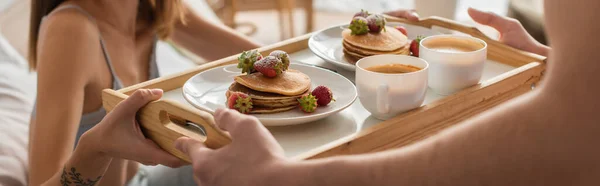 Cropped view of blurred man holding tray with coffee, pancakes and fresh strawberries near woman in bedroom, banner — стоковое фото