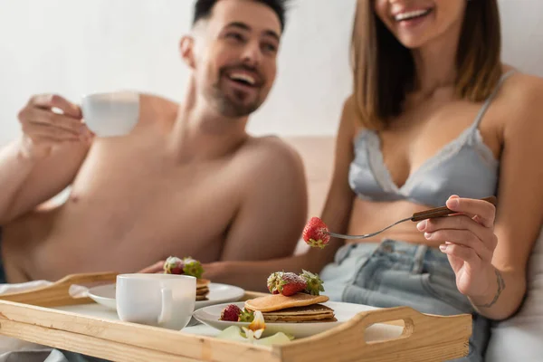 Enfoque selectivo de bandeja con panqueques y café cerca de sexy pareja joven sonriendo sobre fondo borroso - foto de stock