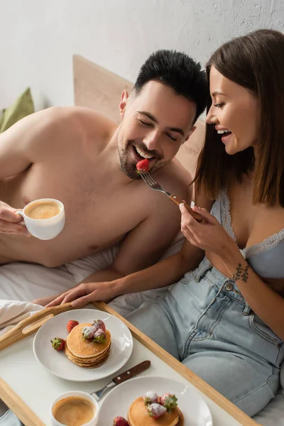 Cheerful woman in jeans and bra feeding boyfriend with strawberries during breakfast in bedroom — Stockfoto