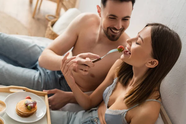 Cheerful shirtless man feeding sexy girlfriend with fresh strawberry during breakfast in bedroom — Foto stock