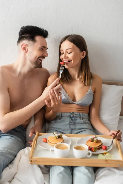 Sexy woman with closed eyes near shirtless man feeding her with strawberry during breakfast in bed — Fotografia de Stock