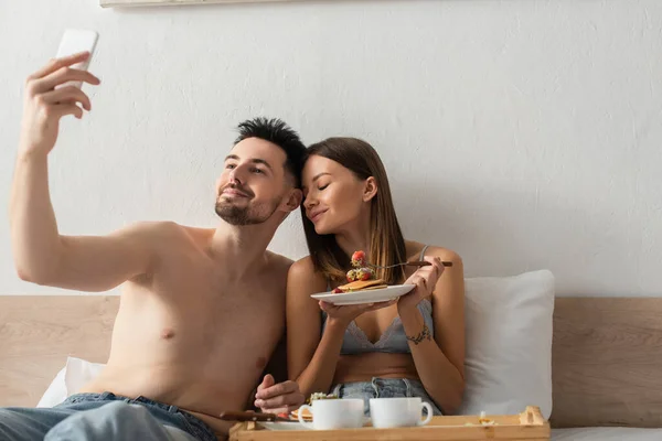 Shirtless man taking selfie with pleased girlfriend eating delicious breakfast in bed - foto de stock