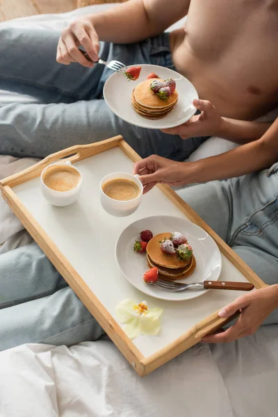 Vista recortada de pareja joven comiendo deliciosos panqueques con fresas frescas para el desayuno en el dormitorio - foto de stock