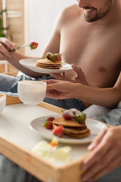 Vista parcial de sonriente hombre sin camisa comiendo sabrosos panqueques con fresas cerca de novia borrosa en el dormitorio - foto de stock