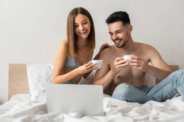 Cheerful couple with coffee cups watching film on computer in bedroom — стоковое фото