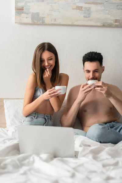Щаслива і сексуальна пара п'є ранкову каву під час перегляду фільму на ноутбуці в спальні — стокове фото