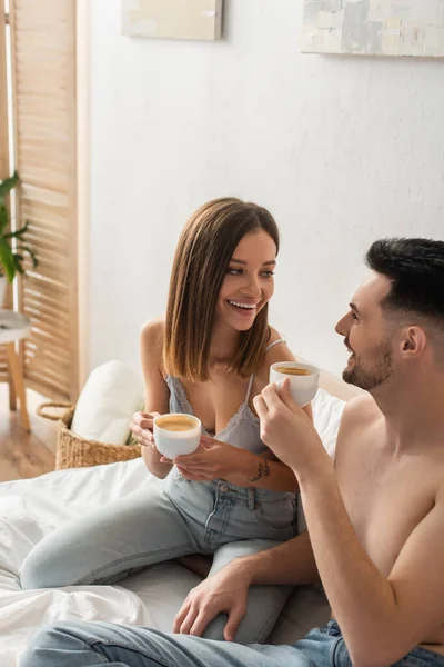 Sexy woman and shirtless man smiling at each other while drinking coffee in bedroom — Stock Photo