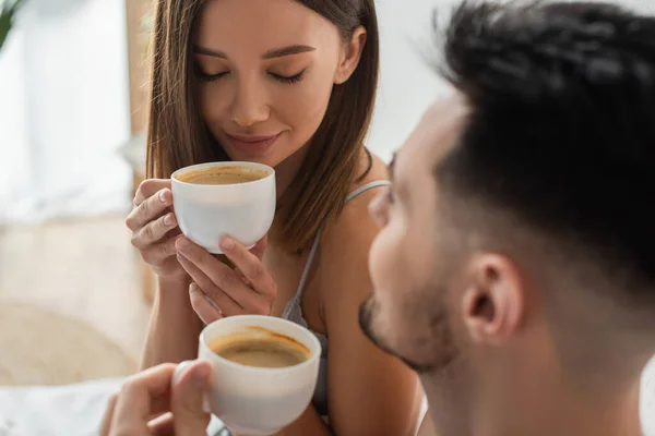 Сексуальна жінка з закритими очима насолоджується ранковою кавою біля розмитого хлопця в спальні — стокове фото