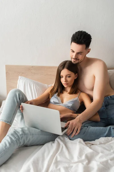Shirtless man and sexy woman in bra and jeans watching film on laptop in bedroom - foto de stock
