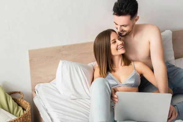 Shirtless man and happy woman in bra looking at each other near laptop in bedroom — стоковое фото