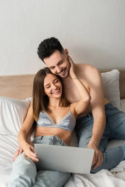 Shirtless man and woman in jeans and bra laughing while watching movie on laptop in bed — стоковое фото