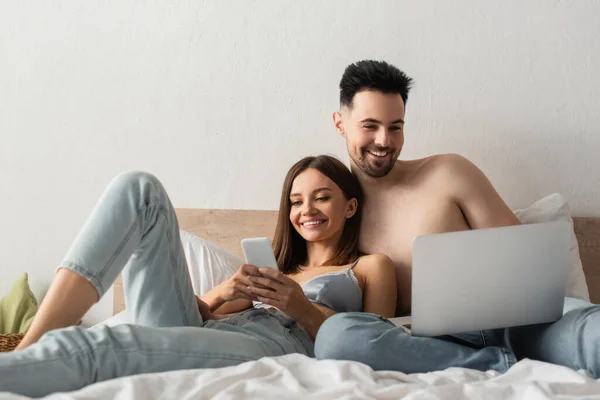 Задоволена пара посміхається, використовуючи гаджети в спальні вдома — стокове фото