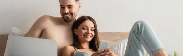 Joyful young lovers using laptop and mobile phone in bedroom, banner — стоковое фото