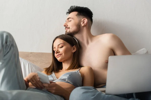 Hombre sin camisa y mujer sexy en sujetador y jeans que sostienen gadgets mientras se relaja con los ojos cerrados en el dormitorio - foto de stock