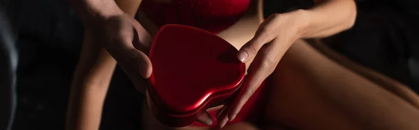 Cropped view of man presenting red heart-shaped gift box to woman on valentines day, banner — Stockfoto