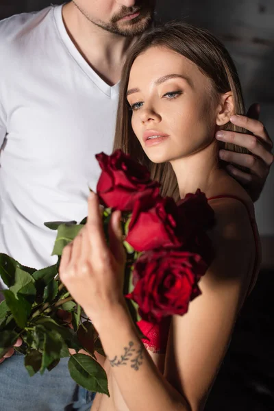 Man in white t-shirt touching woman with red roses in dark bedroom — Stock Photo