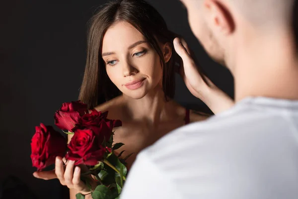 Mujer sensual feliz sosteniendo ramo de rosas rojas cerca de hombre borroso sobre fondo oscuro - foto de stock