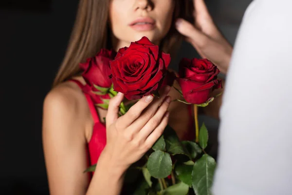 Vista recortada de la mujer borrosa sosteniendo rosas rojas cerca del hombre sobre fondo oscuro - foto de stock
