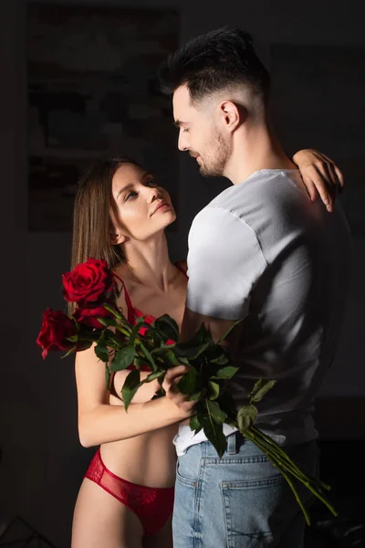 Joven hombre y sexy mujer en lencería mirando unos a otros cerca de rosas rojas en dormitorio oscuro - foto de stock