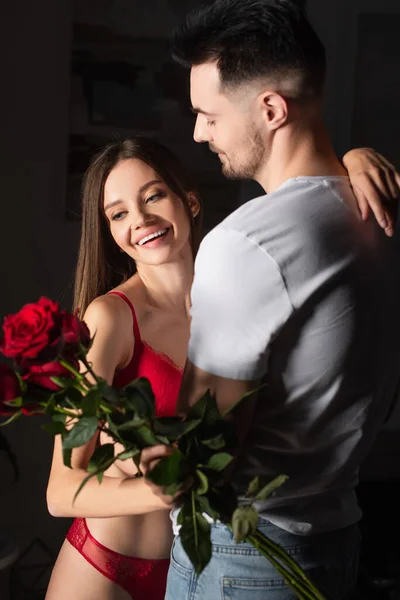 Hombre en camiseta blanca cerca de mujer feliz en ropa interior sexy sosteniendo rosas rojas - foto de stock
