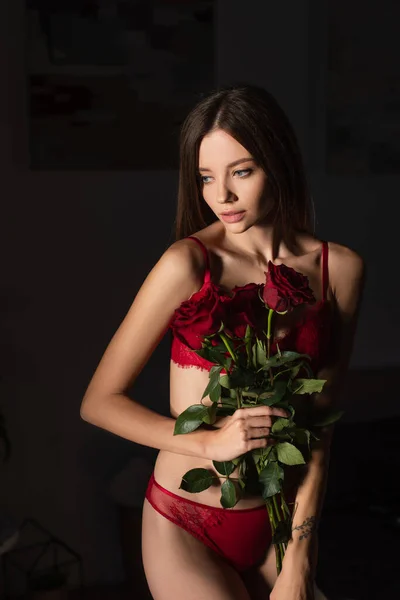 Young sensual woman in red underwear holding red roses in dark bedroom — стоковое фото