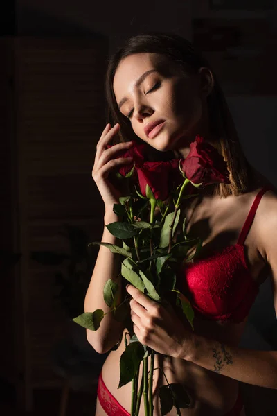 Passionate woman in red underwear standing with closed eyes and roses on dark background - foto de stock