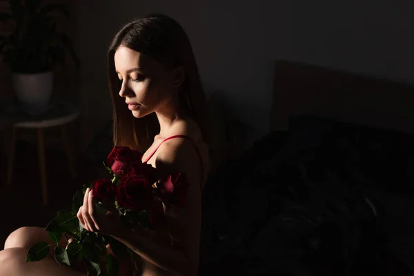 Seductive woman sitting in dark bedroom with bouquet of red roses — Fotografia de Stock