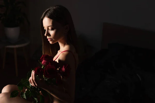 Seductive woman sitting in dark bedroom with bouquet of red roses — Stock Photo