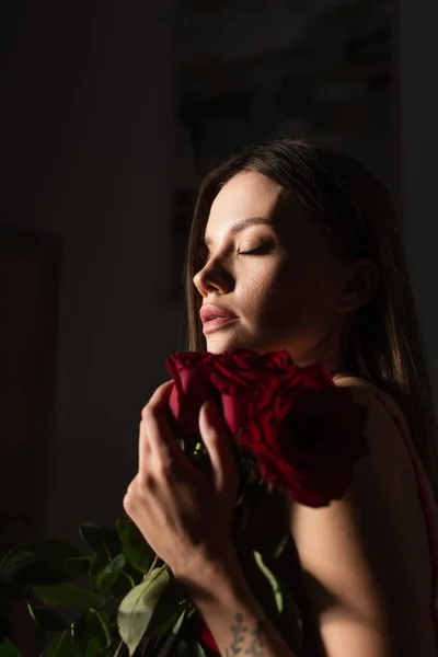 Молода і чуттєва жінка з закритими очима тримає червоні троянди на темному фоні — стокове фото