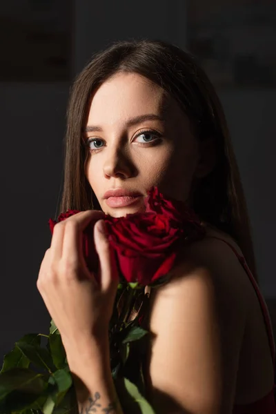 Bonita mujer joven con ramo de rosas rojas mirando a la cámara sobre fondo oscuro - foto de stock