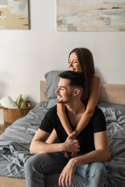 Mujer alegre sonriendo con los ojos cerrados mientras abraza al hombre sentado en la cama y sosteniendo sus manos - foto de stock