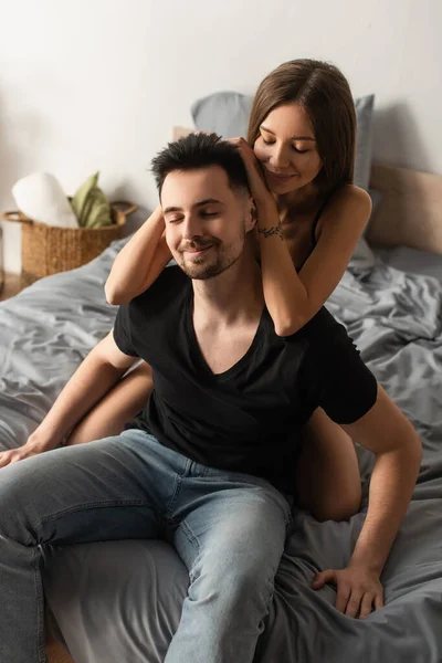 Щаслива жінка обіймає голову усміхненого чоловіка, що сидить на ліжку із закритими очима — стокове фото