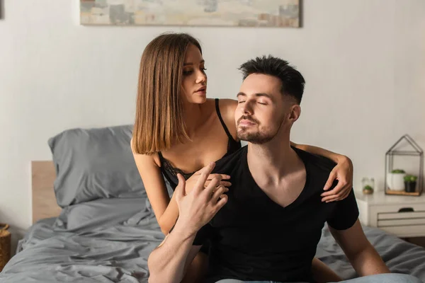 Sensual joven mujer abrazando hombre en negro camiseta sentado en la cama con los ojos cerrados - foto de stock