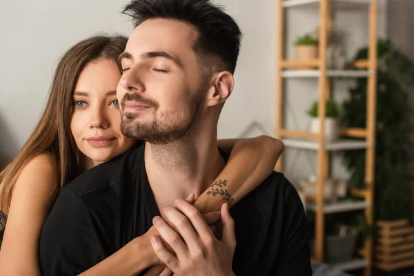 Happy tattooed woman hugging boyfriend in black t-shirt sitting with closed eyes in bedroom — Stockfoto