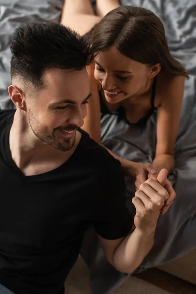 Sexy woman lying on bed and holding hands of happy man in black t-shirt — Stock Photo