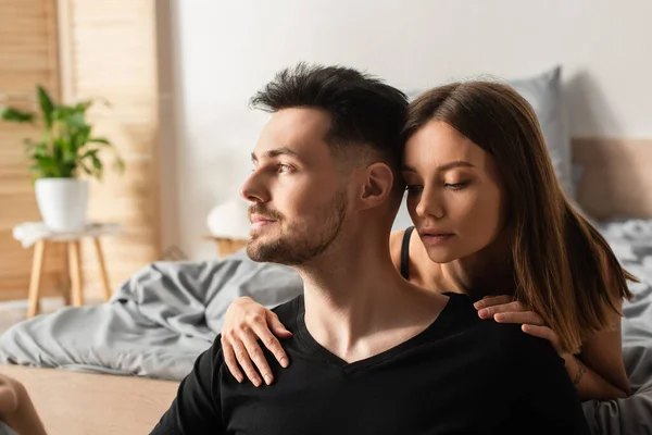 Sensual young woman hugging shoulders of man in black t-shirt looking away in bedroom — стоковое фото