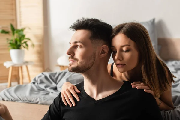 Sensual young woman hugging shoulders of man in black t-shirt looking away in bedroom — Stock Photo