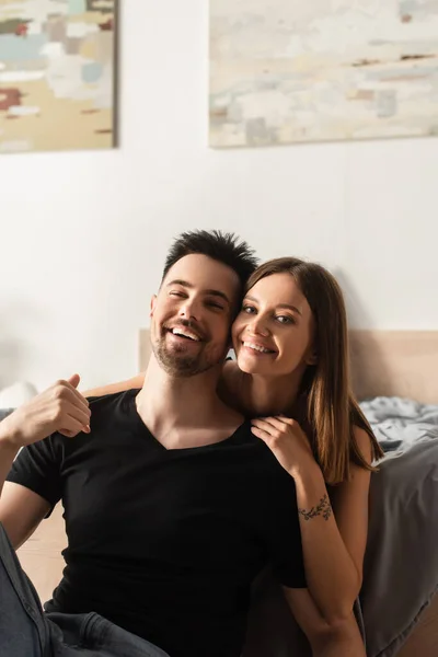 Pretty tattooed woman and man in white t-shirt laughing at camera in bedroom — Stock Photo
